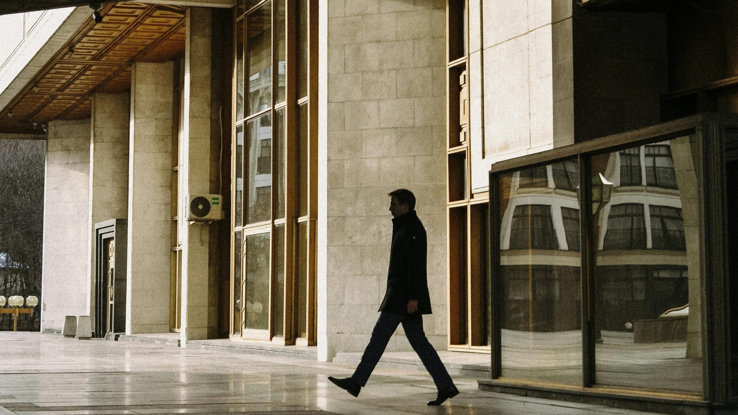 Man in a dark suit walking past the entrance of a large concrete and glass corporate building