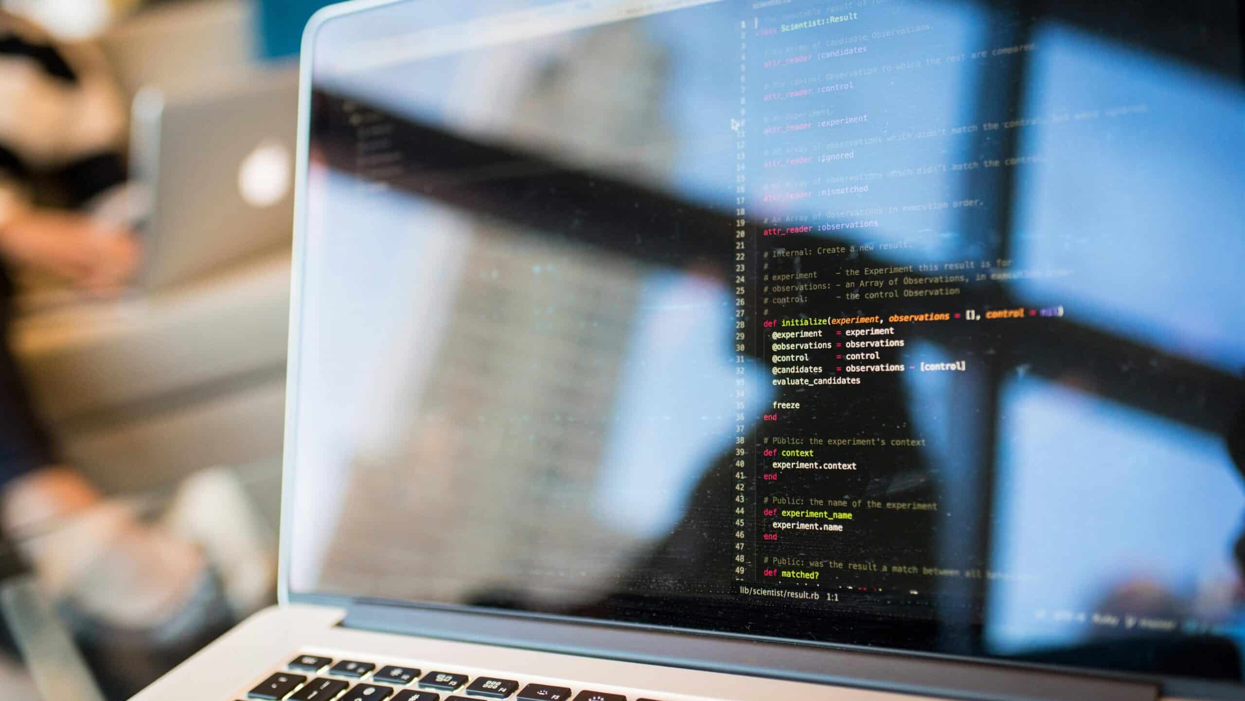 Developer's hands at a laptop with colorful syntax-highlighted code displayed on a monitor
